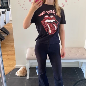 Svart Rolling Stones t-shirt med leopardmönster - Säljer en svart t-shirt med det ikoniska Rolling Stones-trycket. T-shirten har en cool leopardmönstrad tunga i rött och svart, och texten 'The Rolling Stones' i röd stil. Perfekt för en rockig look!
