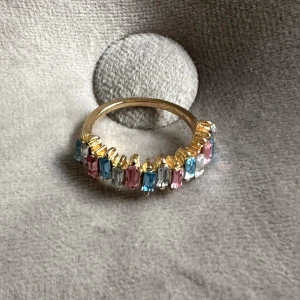 Färgglad ring med stenar - Säljer en superfin ring i guld med färgglada stenar. Den har en rad av rektangulära stenar i blått, rosa och klart, vilket ger en härlig regnbågseffekt. Perfekt för att liva upp vilken outfit som helst!
