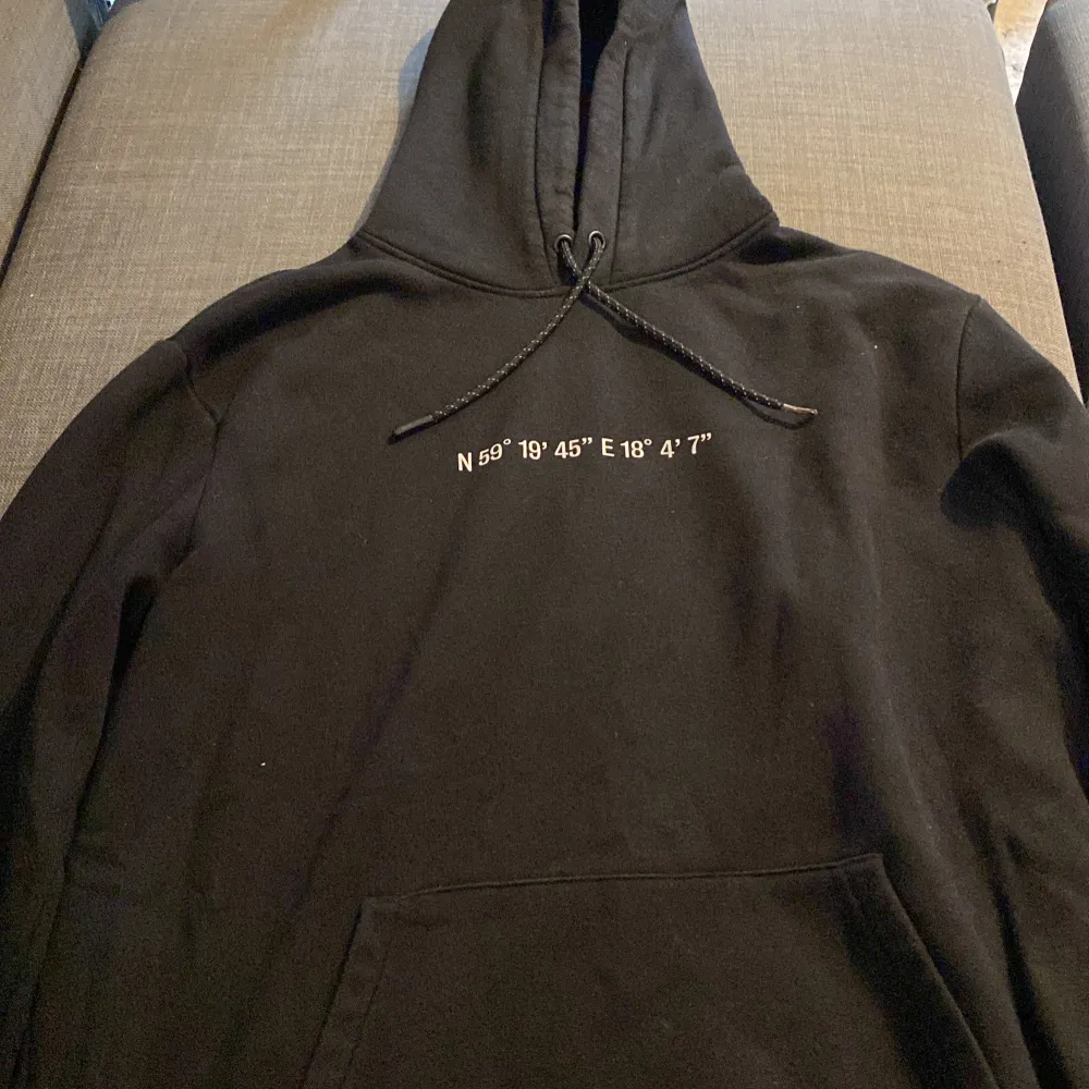 Säljer en svart hoodie med coola textdetaljer både fram och bak. På framsidan finns koordinater tryckta och på baksidan står det 'FUTURE PROOF WORK WEAR TRUE TO REALITY' samt 'ALL SEASON'. Perfekt för alla årstider och superbekväm med en känguruficka framtill. Passar perfekt för en avslappnad stil!. Tröjor & Koftor.