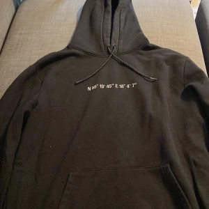 Svart hoodie med textdetaljer - Säljer en svart hoodie med coola textdetaljer både fram och bak. På framsidan finns koordinater tryckta och på baksidan står det 'FUTURE PROOF WORK WEAR TRUE TO REALITY' samt 'ALL SEASON'. Perfekt för alla årstider och superbekväm med en känguruficka framtill. Passar perfekt för en avslappnad stil!