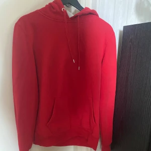 Röd hoodie - Säljer en snygg röd hoodie som är perfekt för kyliga dagar. Den har en klassisk design med en stor ficka framtill och justerbar huva med dragsko. Materialet känns mjukt och bekvämt, perfekt för både vardag och avslappnade tillfällen.