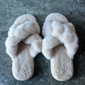 Fluffiga tofflor från Hunkemöller - Säljer ett par supermysiga och fluffiga tofflor från Hunkemöller. De är i en ljus beige färg och har en flätad design på ovansidan. Perfekta för att hålla fötterna varma och bekväma hemma. Sulan har ett mönster med små rosetter för extra grepp.