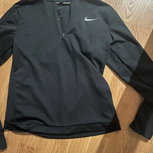 Svart långärmad tröja från Nike - Säljer en svart långärmad tröja från Nike med dragkedja vid halsen. Tröjan är tillverkad i Dri-FIT-material, perfekt för löpning och träning. Den har en stilren design med Nike-logga på bröstet.