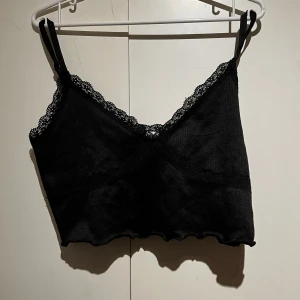 Svart ribbad linnetopp med spetsdetaljer - Snygg svart ribbad linnetopp med spetsdetaljer längs kanterna. Toppen har smala axelband och en v-ringad design som ger en feminin touch. Perfekt för att styla med olika outfits.