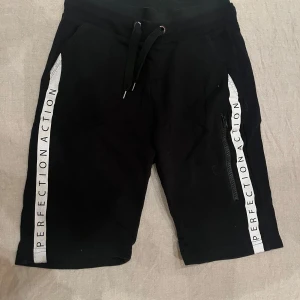 Svarta shorts från Wox 720 - Snygga svarta shorts från Wox 720 med texten 'PERFECTION ACTION' längs sidorna. De har en bekväm dragsko i midjan och praktiska fickor med dragkedja. Perfekta för en avslappnad stil.