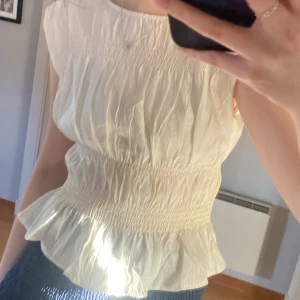 Vit blus - Säljer en söt vit ärmlös blus med smockdetaljer i midjan och volang nedtill. Perfekt för en somrig look. Blusen har en luftig och lätt design som passar bra till både kjol och byxor. Aldrig använd.