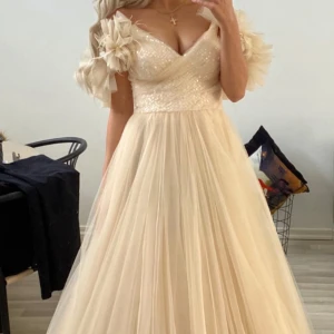 Champagnefärgad balklänning  - Elegant champagnefärgad balklänning med volangdetaljer på axlarna och en v-ringad överdel. Klänningen har en lång, flödande kjol i tyll och en glittrig överdel med dragkedja i ryggen. Köpt på edessas mode i Södertälje. Använd en gång av en annan som jag köpte klänningen av. Jag har aldrig använt den. 