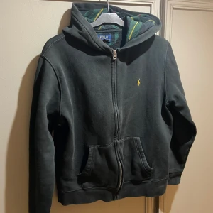 Svart Zip hoodie från Ralph Lauren - Säljer en svart hoodie från Ralph Lauren med dragkedja och huva. Storlek 170. Den är i fint skick och prisvärd. Passar inte priset så kom med prisförslag så tar vi de där! OBS STORLEK I ANNONS STÄMMER EJ!