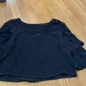 Svart blus från H&M - Säljer en elegant svart blus från H&M med vackra spetsdetaljer vid halsen. Blusen har korta ärmar med volang och en luftig passform. Perfekt för en stilren look.