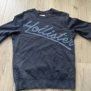 Mörkgrå tröja från Hollister - Säljer en snygg mörkgrå tröja från Hollister med deras logga i stort tryck över bröstet. Tröjan har långa ärmar och en klassisk rund halsringning. Perfekt för en avslappnad stil.