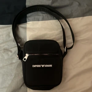 Svart axelväska från Emporio Armani - Snygg svart axelväska från Emporio Armani med justerbar rem och dragkedja. Perfekt för att bära dina nödvändigheter med stil. Väskan har en minimalistisk design med logotyp framtill.