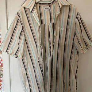 Randig kortärmad skjorta från Weekday - Snygg randig oversize skjorta från Weekday i beige, svart och mintgrön. Skjortan har korta ärmar och en klassisk krage. Perfekt för en avslappnad stil.