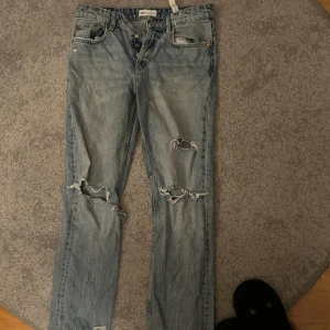 Blå jeans från Zara - Säljer ett par blå jeans från Zara med slitna detaljer och knappgylf. De har en klassisk femficksdesign och är perfekta för en avslappnad stil.