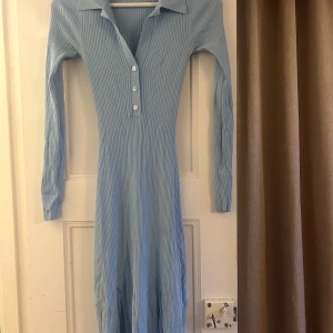 Ljusblå ribbad klänning från ADOORE - Säljer en ljusblå ribbad klänning från ADOORE med långa ärmar och knappar framtill. Klänningen har en klassisk krage och är i ett mjukt material. Den har två mindre hål i bak, som går att laga. 
