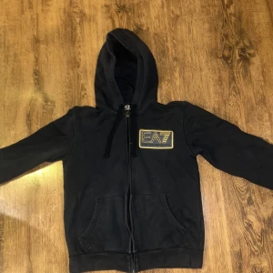 Svart hoodie från Emporio Armani - Säljer en stilren svart hoodie från Emporio Armani med guldtryck på bröstet. Den har dragkedja och en praktisk huva. Perfekt för en avslappnad look.