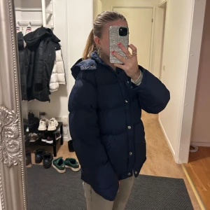 Mörkblå dunjacka med huva - Säljer en mysig mörkblå dunjacka med luva och dragkedja. Jackan har en pufferdesign och knappar framtill. Perfekt för kalla dagar!