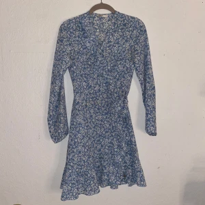 Blommig blå klänning - Säljer en söt blå klänning med blommigt mönster. Klänningen har långa ärmar och en knytning på sidan. Perfekt för vår och sommar!