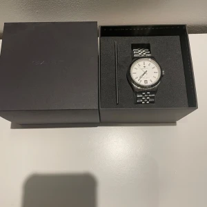 Silverfärgad klocka från GANT (Nypris 2300) - Elegant silverfärgad klocka från GANT med vit urtavla och datumvisning. Klockan har en klassisk länkarmband i rostfritt stål och levereras i originalförpackning med extra länkbitar för justering. Perfekt för den stilmedvetna!