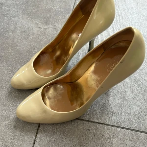 Beige pumps från Guess - Eleganta beige pumps från Guess med hög klack och spetsig tå. Skorna har en glansig yta som ger en lyxig känsla. Perfekta för att lyfta din outfit till nästa nivå.