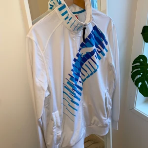 Vit tröja från Puma med blått mönster - Säljer en vit tröja från Puma med ett coolt blått mönster över bröstet. Jackan har dragkedja och långa ärmar, perfekt för en sportig look. Den har också praktiska fickor på sidorna.