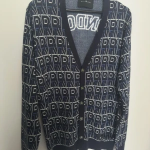 Mönstrad kofta från Richmond - Snygg kofta från Richmond med ett unikt mönster i blått och vitt. Koftan har långa ärmar och knappar framtill. Texten 'LONDON' pryder ryggen, vilket ger en cool touch. Perfekt för dig som vill sticka ut!