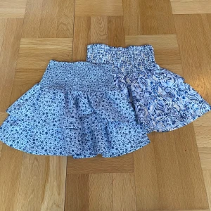 Två blommiga kortkjolar - Säljer två söta kortkjolar med blommigt mönster. Den ena är ljusblå med små blommor och den andra är vit med blå beaga mönster. Båda har en elastisk midja för bekväm passform. Perfekta för sommarens varma dagar!