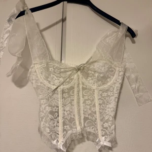 Vit spetskorsett med knytband - Elegant vit korsett i spets med knytband framtill och dekorativa rosetter. Axelbanden är justerbara och ger en romantisk look. Perfekt för speciella tillfällen.
