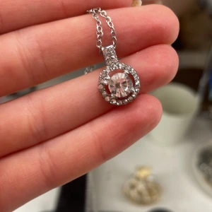 Silverfärgat halsband med glittrig berlock - Elegant silverfärgat halsband med en rund berlock prydd med gnistrande stenar. Perfekt för att ge en touch av glamour till din outfit. Kedjan är tunn och smidig med ett enkelt lås.