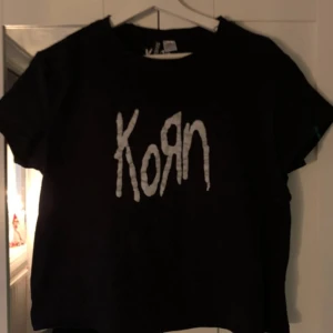 Svart t-shirt med Korn-tryck - Snygg svart t-shirt med det ikoniska Korn-trycket i vitt på framsidan. Perfekt för fans av bandet som vill visa sin stil. T-shirten har en klassisk passform och korta ärmar.