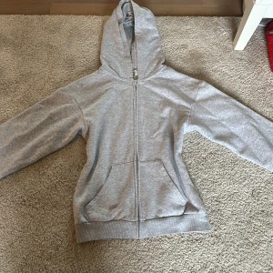Grå zip-up hoodie från H&M - Säljer en klassisk grå hoodie från H&M med dragkedja och fickor framtill. Perfekt för en avslappnad stil. Den har en bekväm passform och är tillverkad i mjukt material.