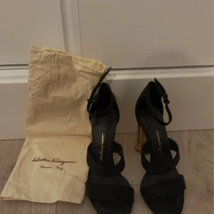 Svarta klackskor från Salvatore Ferragamo - Eleganta svarta klackskor från Salvatore Ferragamo med unika guldfärgade klackar. Skorna har en stilren design med remmar och en justerbar spänne runt ankeln. Perfekta för att ge en lyxig touch till din outfit.