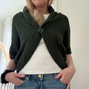 Mörkgrön stickad bolerotopp - Säljer en mysig mörkgrön stickad bolerotopp med en stor knapp framtill. Den har trekvartsärmar och en ribbad kant som ger en snygg passform. 