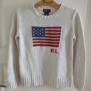 Vit stickad USA-tröja Ralph Lauren - Säljer ett riktigt snyggt exemplar av superpopulära stickade tröjan från Ralph Lauren. Märkt stl 10-12år, sitter ungefär som en dam stl XXS eller möjligen XS, se mått. Mycket fint skick, inga defekter! Mått: Armhåla till armhåla 42cm, bredd längst ner 40cm, längd från mitt på axeln 55cm.