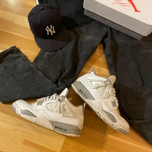 Jordan 4 oreo - Snygga Nike Air Jordans i vitt och grått med klassisk design. Skorna har snörning och en ikonisk Jumpman-logga i rött på tungan och sulan. Perfekta för sneaker-entusiaster som vill ha en stilren look.