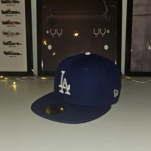 Mörk blå keps från New Era - Snygg mörk blå keps från New Era med LA-logga framtill. Kepsen är en 59FIFTY-modell och är tillverkad i 100% polyester. Perfekt för sportiga tillfällen eller som en stilren accessoar. Använd max 4 gånger.