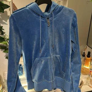 Blå velour hoodie från Juicy Couture - Säljer en mjuk och bekväm blå velour hoodie från Juicy Couture. Den har en dragkedja framtill och en praktisk känguruficka. Perfekt för en avslappnad stil med en touch av lyx. 💙.   NYPRIS 1 400 kr säljer för 399 