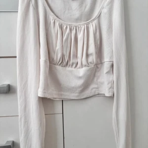vit ribbad långärmad topp - Säljer en stilren beige ribbad topp med långa ärmar. Toppen har en rundad halsringning och en lätt rynkad detalj framtill som ger en fin passform. Perfekt för en avslappnad look.