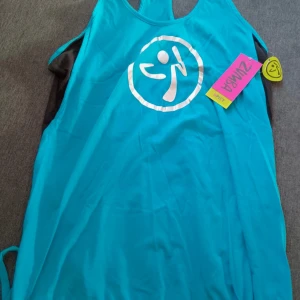 Turkos linne från Zumba - Säljer ett turkost linne från Zumba med ett stort tryck framtill. Linnet är tillverkat i en blandning av bomull och polyester, vilket gör det bekvämt och lätt. Perfekt för dans eller träning! Ny med prislapp.