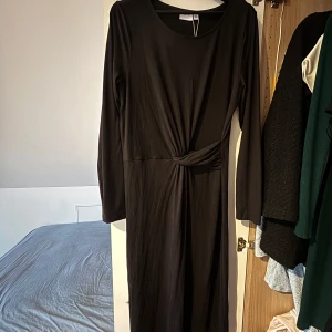 Svart klänning från Vila - Elegant svart klänning från Vila med långärmad design och en stilren knutdetalj i midjan. Perfekt för alla tillfällen. Klänningen har en rund halsringning och är tillverkad i ett mjukt material som ger en bekväm passform. Passar M. Prislapp finns kvar