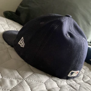 Mörkblå keps från New Era - Snygg mörkblå keps från New Era med ett broderat rött 'B' framtill. Perfekt för sportfantaster och vardagsbruk. Kepsen har en klassisk design och är tillverkad i bomull för en bekväm passform.