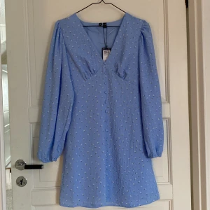 Blå blommig klänning från Vero Moda - Säljer en söt blå klänning från Vero Moda med små blommor och v-ringning. Klänningen har långa ärmar och knappar framtill. Perfekt för en somrig look! 🌸 aldrig använd 