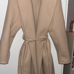 Beige kappa med knytbälte - Säljer en stilren beige kappa med knytbälte i midjan. Den har långa ärmar och en klassisk design som passar perfekt för kyligare dagar. Perfekt för att hålla stilen i höst och vinter.