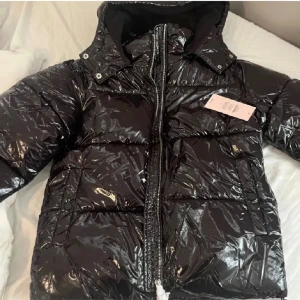 Svart pufferjacka från Nelly - Snygg svart pufferjacka från Nelly med glansig finish. Jackan har en dragkedja framtill och en hög krage med huva. Perfekt för kyliga dagar när du vill hålla stilen. 🖤