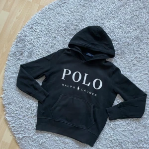 Svart hoodie från Ralph Lauren - Säljer en svart hoodie från Ralph Lauren med stor POLO-logga på framsidan. Tröjan har en klassisk känguruficka och en bekväm huva. Perfekt för en avslappnad stil.