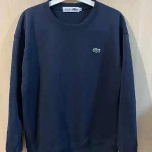 Mörkblå tröja från Lacoste - Säljer en stilren mörkblå tröja från Lacoste med det klassiska krokodilmärket på bröstet. Tröjan har rund hals och långa ärmar, perfekt för en avslappnad look. Passar bra till både jeans och chinos. 🐊