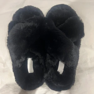 Svarta fluffiga tofflor - Mysiga svarta tofflor med fluffigt pälsmaterial. De har en enkel design med öppen tå och korsade remmar. Aldrig använda då det inte passade mig.
