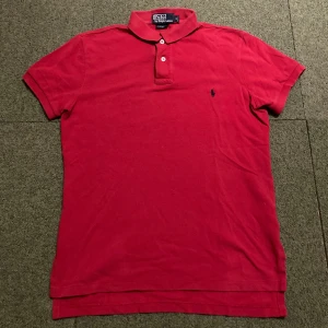 Röd pikétröja från Ralph Lauren - Säljer en klassisk röd pikétröja från Ralph Lauren med den ikoniska broderade loggan på bröstet. Tröjan har korta ärmar och en stilren krage med knappar. Perfekt för en avslappnad och stilren look.