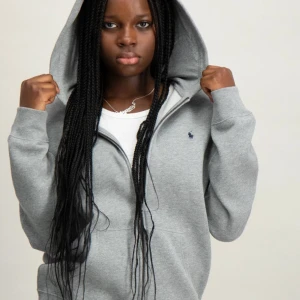 Grå hoodie från Ralph Lauren - Säljer en stilren grå hoodie från Ralph Lauren med dragkedja och huva. Tröjan har en liten broderad logotyp på bröstet och en praktisk ficka framtill. Perfekt för en avslappnad look. Säljer då vi råkade köpa barnstorlek
