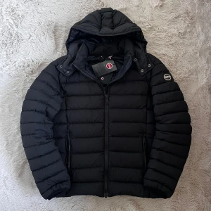 Svart pufferjacka från Colmar - Säljer en svart pufferjacka från Colmar med huva och dragkedja. Jackan har en stilren design med quiltade paneler och är perfekt för kyligare väder. Den har en praktisk dragkedja framtill och två sidofickor med dragkedjor. St 50 motsvarande M 