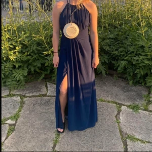 Mörkblå balklänning med slits - Elegant mörkblå långklänning med halterneck och hög slits. Perfekt för en sommarkväll eller bal. Klänningen har en smickrande passform och ger en stilren look.
